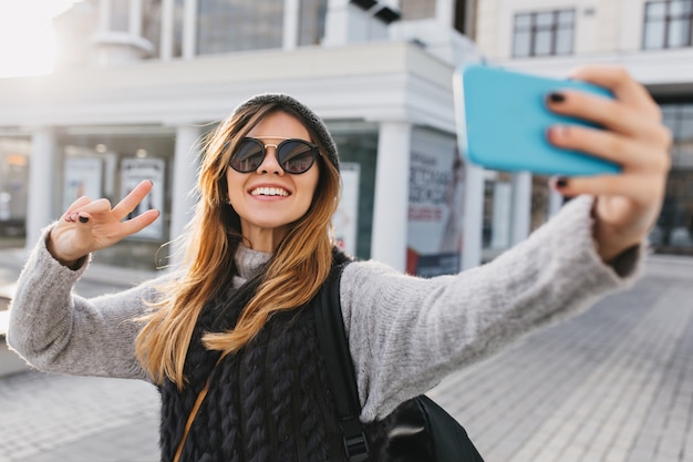 Бесплатное фото Модная очаровательная красивая женщина в современных солнцезащитных очках, теплый зимний свитер делает селфи-портрет на улице в центре города. стильно выглядеть, веселиться, выражать позитивные яркие эмоции.