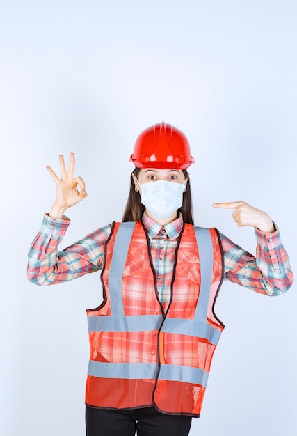 Free photo female construction engineer in red helmet and safety mask showing successful hand sign