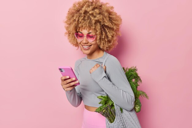 Бесплатное фото Женщина-вегетарианка с кудрявыми волосами носит сумку из органической ткани со свежими зелеными овощами, использует смартфон, просматривает меню, придерживается диеты, носит сердечки и спортивную одежду, изолированную на розовом фоне