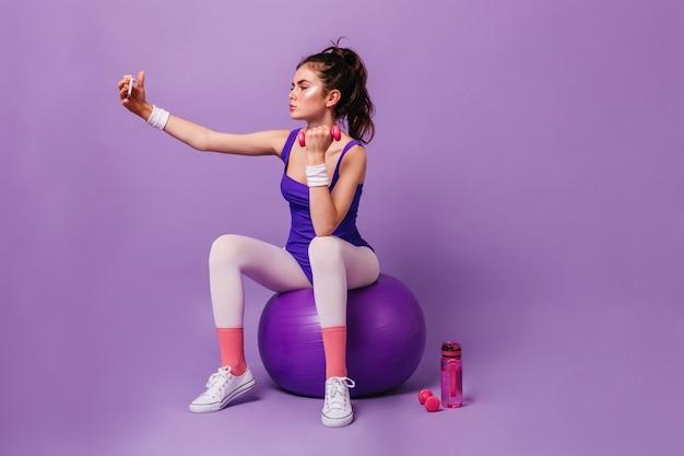 Бесплатное фото Женщина-фитнес-тренер сидит на спортивном мяче, делает упражнения с гантелями и делает селфи на фиолетовой стене