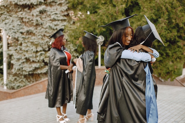 Сосредоточьтесь на двух молодых афроамериканских студентках, одетых в черное выпускное платье. Кампус как фон. Девочки обнимаются