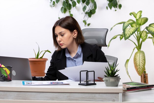 A front view beautiful young businesswoman in black jacket and blue shirt working with laptop and documents in front of table business job office