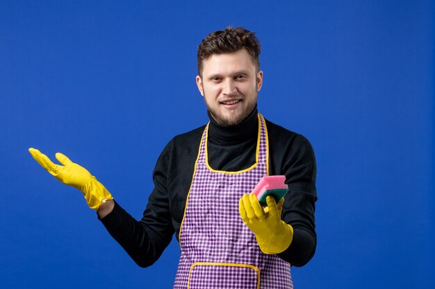 Front view of smiling male housekeeper holding up sponge on blue wall