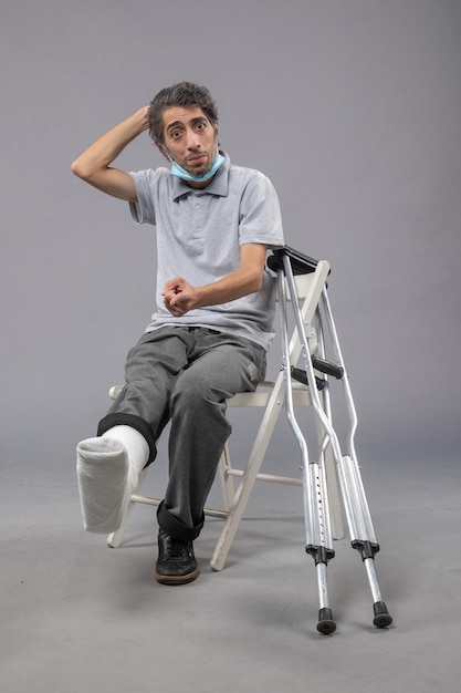 Бесплатное фото Вид спереди молодого мужчины, сидящего со сломанной ногой и связанной повязкой на серой стене, боль в ноге, скручивание стопы, мужчина, несчастный случай