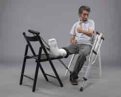 Бесплатное фото Вид спереди молодой мужчина, сидящий со сломанной ногой и костылями, улыбаясь на серой стене, несчастный случай, боль в ноге, сломанная ступня