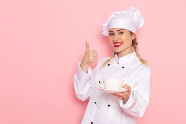 Вид спереди молодая женщина-повар в белом костюме повара держит чашку кофе с легкой улыбкой на розовом космическом поваре