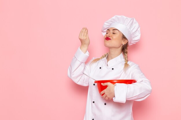 Вид спереди молодая женщина-повар в белом костюме повара держит красную пластиковую миску на розовом космическом поваре