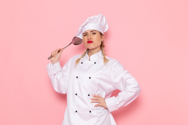 Вид спереди молодая женщина-повар в белом костюме повара держит серебряную ложку на розовом космическом поваре