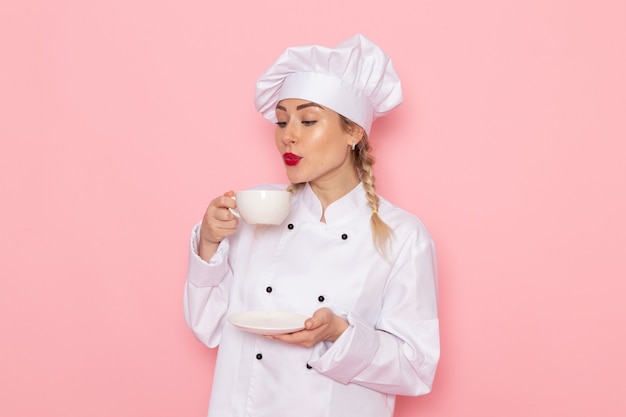 Вид спереди молодая женщина-повар в белом костюме повара держит белую чашку с кофе на розовом космическом поваре