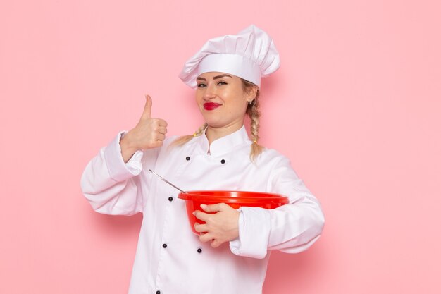 Вид спереди молодая женщина-повар в белом костюме, держащая красную миску, улыбаясь в розовом пространстве, повар, кухня, работа, работа, фото