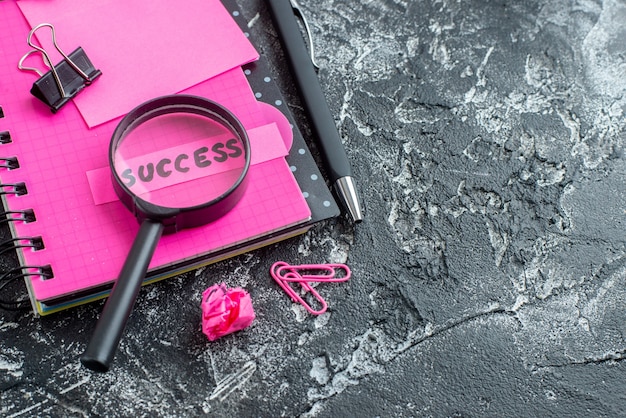 Бесплатное фото Розовый блокнот с ручкой-лупой и заметкой об успехе на сером фоне