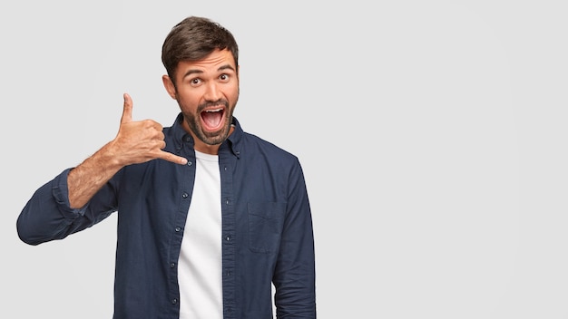 Free photo funny happy male keeps hand near ear, makes call gesture, pretends speaking with someone, keeps mouth widely opened, dressed in fashionable shirt, isolated over white wall, free space aside.