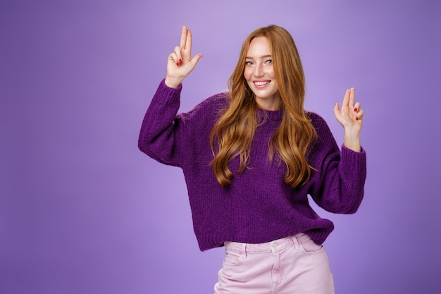 Бесплатное фото Девушка в счастливом и энергичном настроении танцует с удовольствием, делая жест пистолета пальцами, кокетливо улыбаясь в камеру, чувствуя себя взволнованной и радостной на удивительной вечеринке, позирующей в фиолетовом свитере над фиолетовой стеной.