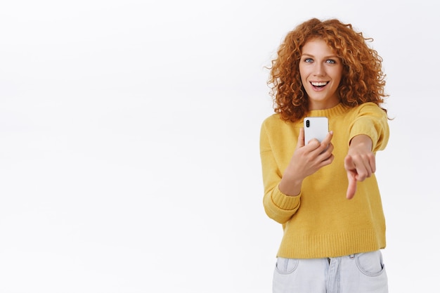 Бесплатное фото Девушка довольна тем, что вы фотографируете на смартфоне. привлекательная эмоциональная рыжая кудрявая женщина держит мобильный телефон, указывая камеру указательным пальцем, радостно улыбаясь, белая стена
