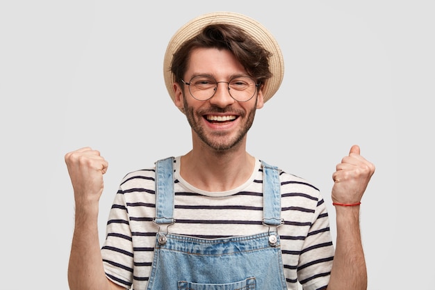 Бесплатное фото Симпатичный позитивный фермер-мужчина поднимает кулаки, чувствует себя довольным и возбужденным, добивается больших успехов в сельскохозяйственной сфере, носит повседневный комбинезон, полосатый свитер, соломенную шляпу, имеет широкую улыбку.