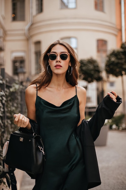 Free photo gorgeous young brunette with red lips and tattoo on body sexy green silk dress black jacket and bag trendy sunglasses standing outdoors against old city architecture