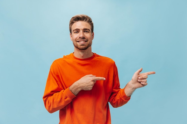 Free photo handsome bearded man with stylish hairstyle in bright orange sweatshirt looking into camera smiling and pointing to place for text