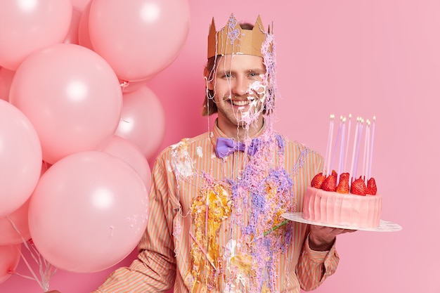 Бесплатное фото Красивый веселый мужчина стоит грязно со сливками и держит вкусный клубничный торт празднует день рождения с праздничными атрибутами, изолированными над розовой стеной