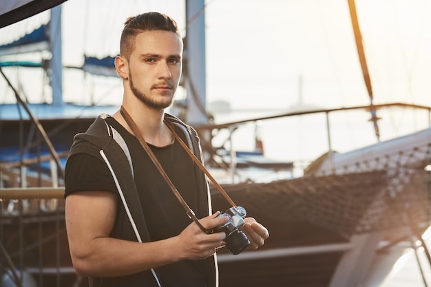 Бесплатное фото Красивый уверенный в себе парень со стильной стрижкой стоит возле крутой яхты, держит фотоаппарат, серьезно смотрит и фокусируется во время фотосессии в гавани, снимает пейзажи
