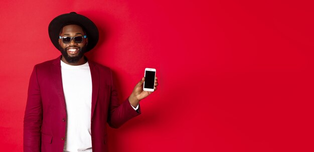 Красивый и стильный темнокожий мужчина показывает экран телефона на камеру, рекомендуя интернет-магазин или приложение