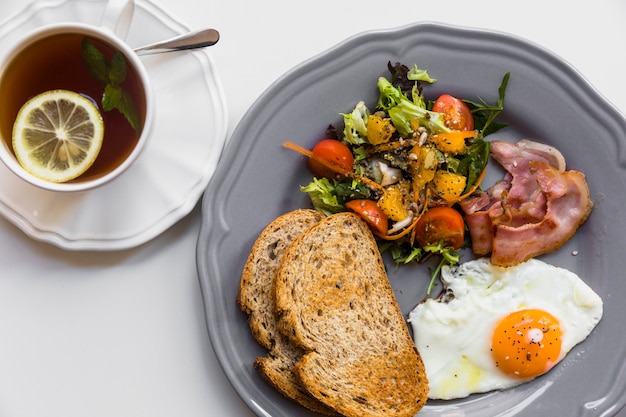 Половину жареного яйца; тост; салат; бекон на серой тарелке с чашкой чая с лимоном и мятой на белом фоне