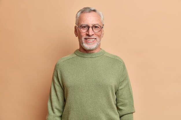 Half length shot of cheerful senior man smiles happily with white teeth wears optical glasses and sweater isolated over brown wall