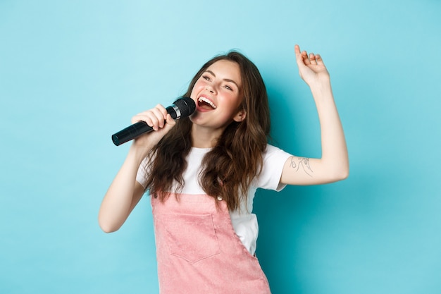 Free photo happy young woman perform song, singer holding microphone, dancing and singing at karaoke, standing over blue background