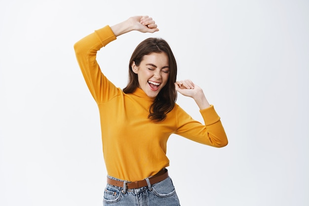 Free photo happy attractive woman dancing and having fun, raising hands up carefree, enjoying music, standing against white wall