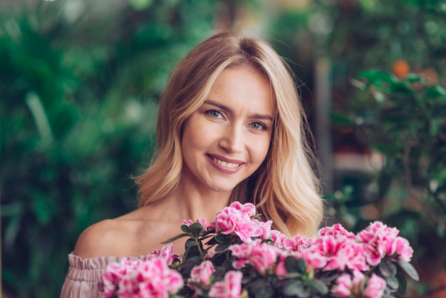 Free photo happy blonde young woman standing behind the pink flowers with blurred background