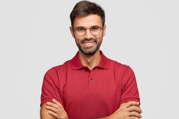 Free photo happy confident male entrepreneur with postive smile, has beard and mustache, keeps arms folded, being in high spirit after successful meeting with partners, poses against white wall, dressed casually