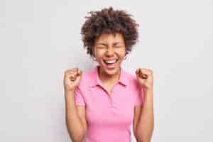 Бесплатное фото Счастливая кудрявая афро-американка накачивает кулаком и радуется положительным результатам в повседневной розовой футболке.