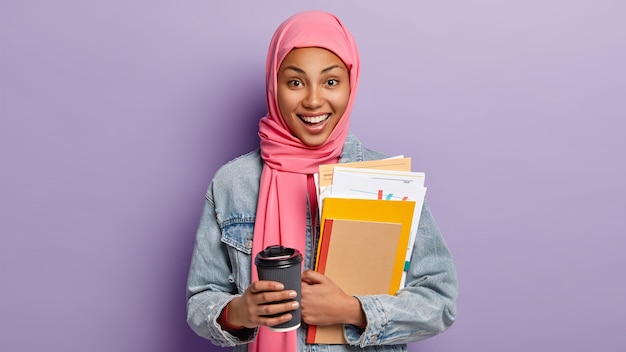 Free photo happy ethnic female student has coffee break, holds takeaway cup of drink, carries notebook and papers, has pink scarf on head, islamic religious views, poses indoor. people, culture, tradition