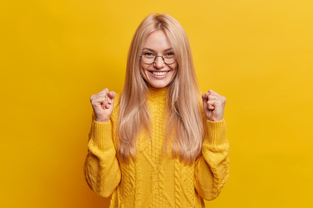 Бесплатное фото Счастливая радостная блондинка сжимает кулаки и празднует хороший результат, широко улыбается, в приподнятом настроении делает торжественный жест, носит очки и зимний свитер