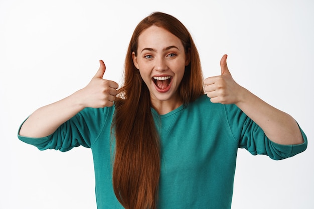 Бесплатное фото Счастливая смеющаяся рыжая девушка, одобрительно показывает большие пальцы вверх, довольная чем-то крутым, хвалит и комплимент, дает положительный отзыв на белом.