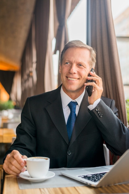 Счастливый зрелый бизнесмен разговаривает по мобильному телефону с чашкой кофе и ноутбук на столе