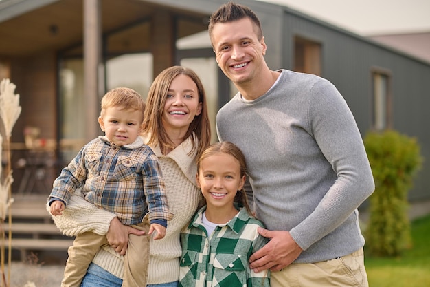 Free photo happy people.. young beautiful woman with little boy attractive man and long-haired school-age girl in casual clothes hugging joyful looking at camera standing near house