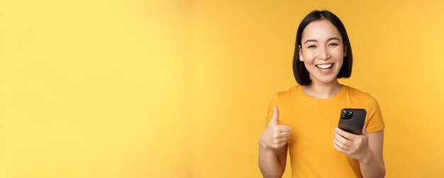 Счастливая улыбающаяся азиатская девушка держит мобильный телефон и показывает палец вверх, рекомендуя приложение на смарт