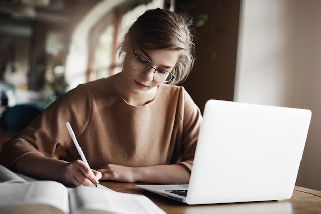 Трудолюбивая сосредоточенная женщина в модных очках, сосредоточенная на написании эссе, сидит в уютном кафе рядом с ноутбуком, работает и тщательно делает заметки.