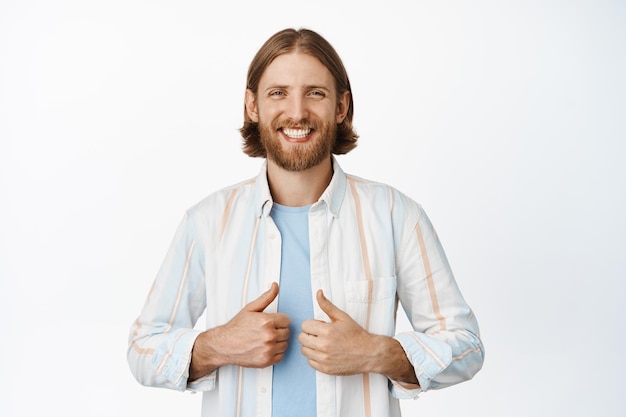 Бесплатное фото Изображение красивого бородатого парня со светлыми волосами, счастливо улыбающегося с белыми зубами, показывающего большой палец вверх, выглядящего уверенным и расслабленным, стоящего на белом фоне.