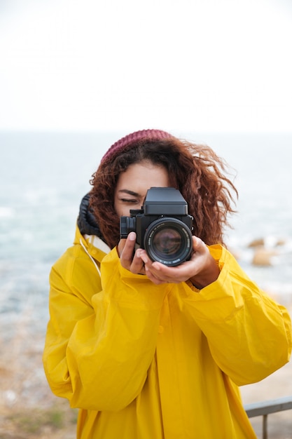 Бесплатное фото Изображение счастливого африканского вьющегося фотографа молодой женщины нося желтое пальто держа камеру и гуляя outdoors