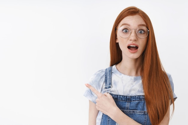 Free photo impressed cute redhead girl with long hair open mouth astonished look camera amused pointing upper left corner questioned gaze you asking question about interesting promo white background