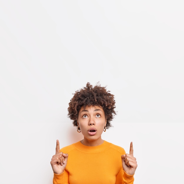 Free photo impressed beautiful afro american woman looks shocked above indicates at copy space shows incredible offer surprised by big sales wears casual clothes isolated over white wall. promotion