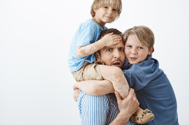 Бесплатное фото Снимок в помещении: обеспокоенный усталый отец держит симпатичного белокурого сына с витилиго на плечах, хмурится и волнуется, а старший брат висит на груди отца