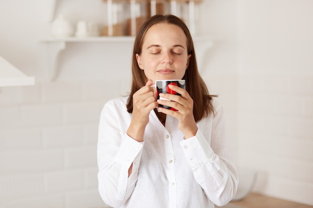 Бесплатное фото Крытый снимок счастливой красивой женщины, наслаждающейся чашкой кофе или чая на кухне утром, пьющей горячий напиток, стоящей с закрытыми глазами с расслабленным выражением лица.