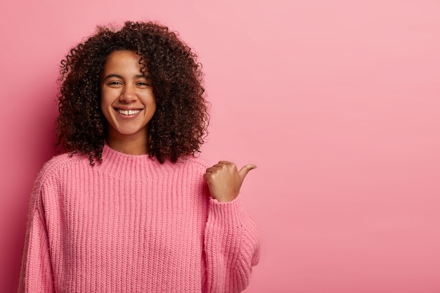 Free photo indoor shot of satisfied afro woman has dark curly hair, points aside with thumb, demonstrates nice copy space for your advertising, has friendly expression, wears oversized pink sweater.