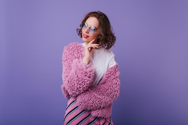 Бесплатное фото Внутренний портрет элегантной кавказской женщины в модной яркой куртке мечтательная девушка в солнечных очках с удовольствием позирует в шубе на фиолетовом фоне