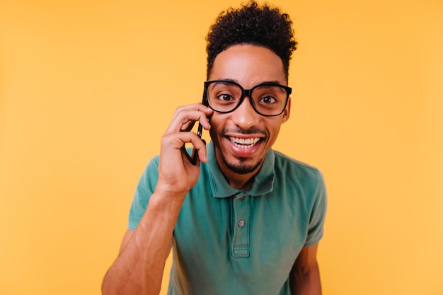 Free photo interested dark-eyed man in glasses talking on phone. happy african guy wears green clothes posing with smartphone.