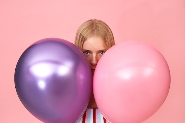 Бесплатное фото Изолированные портрет загадочной молодой блондинки с веснушками и пирсингом на лице позирует на розовом, прячась за двумя блестящими металлическими гелиевыми шарами