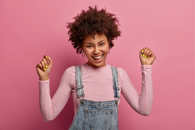 Joyful curly young woman raises arms, makes fit pump, smiles joyfully, dressed in casual wear, being in high spirit, laughs out loudly, poses against pink pastel wall, feels like winner