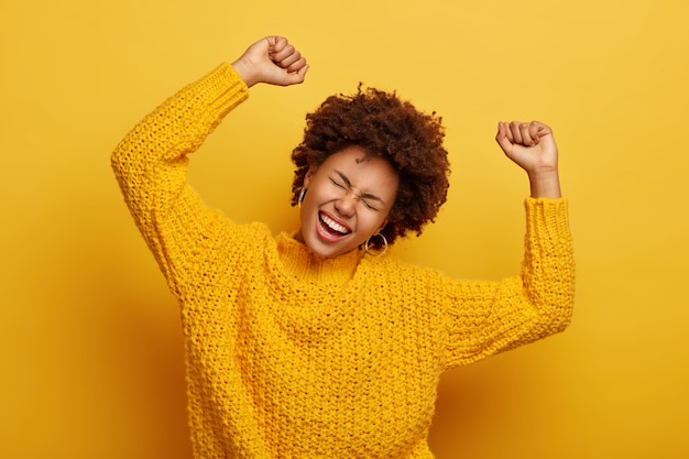 Free photo joyful afro woman raises arms, tilts head, dressed in casual knitted jumper, laughs from happiness, celebrates victory, isolated on yellow
